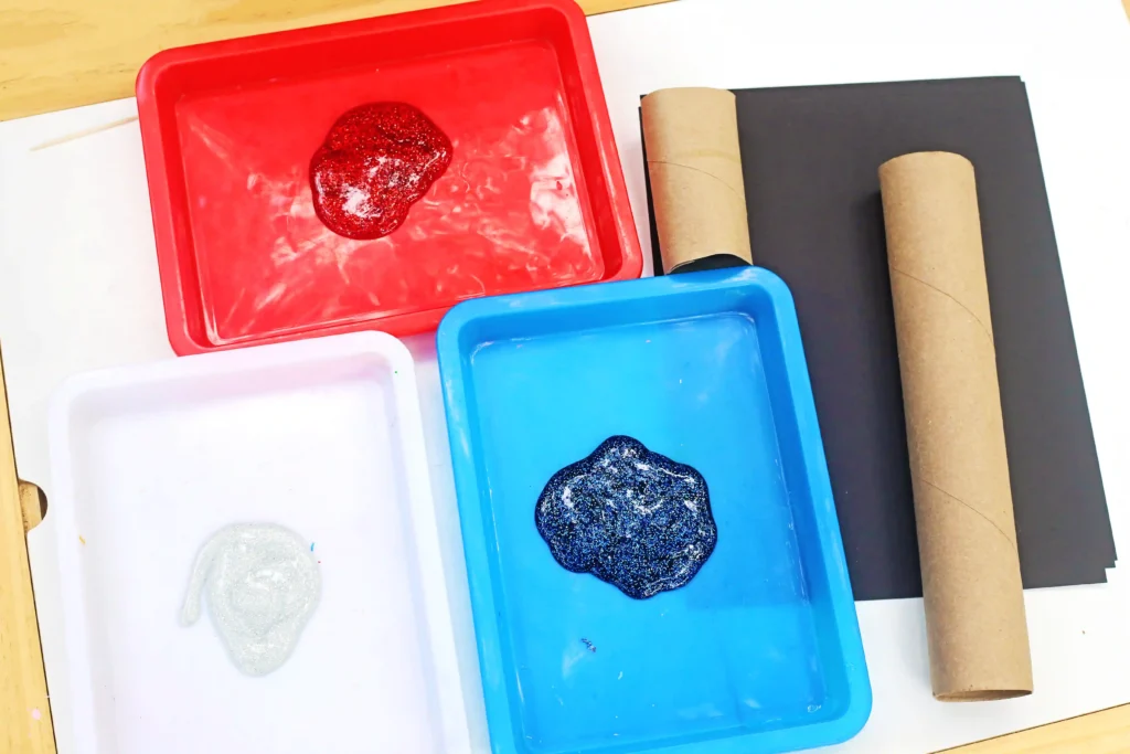 Toilet Paper Roll Firework Craft - Cultivating Brilliant Minds supplies for the toilet paper roll firework craft: Art workspace with red and blue paint trays containing glittery paint, white paper, and several cardboard tubes on a black sheet.