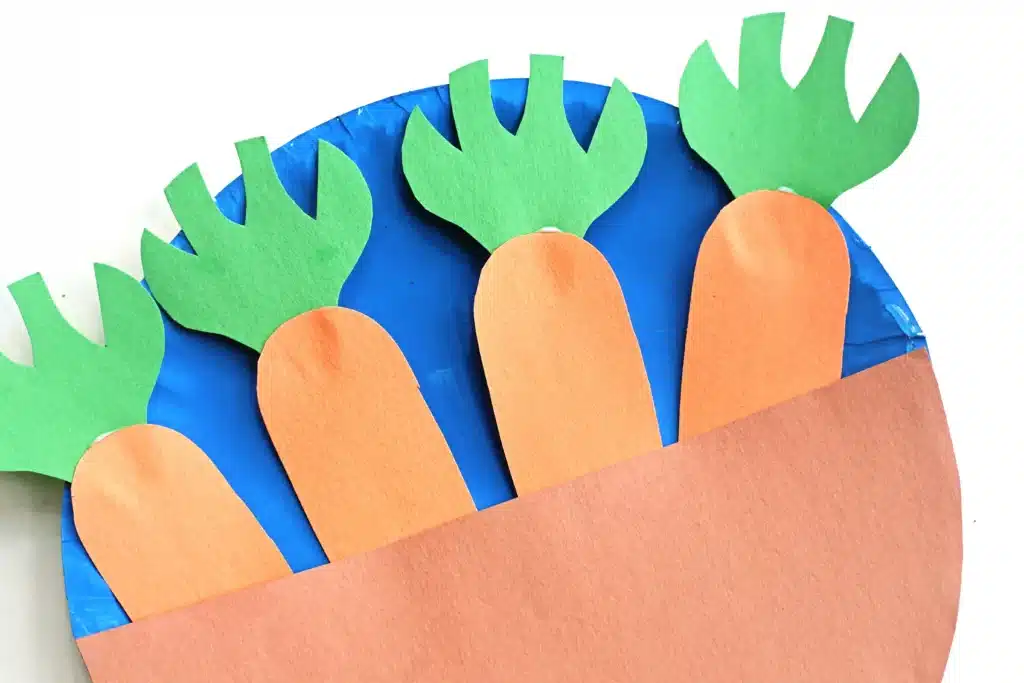Carrot Garden Paper Plate Craft - Cultivating Brilliant Minds a blue paper plate with a brown piece of construction paper glued on with construction paper carrots tucked in