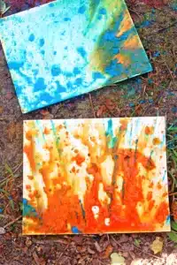 Two abstract painted canvases lying on dirt and fallen leaves, one blue-green with splatters, the other orange and yellow with streaks of color outdoors on the ground.