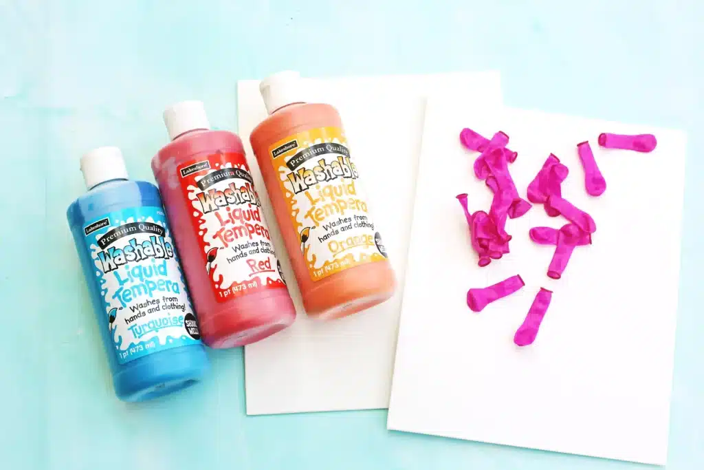 Water Balloon Splatter Art - Cultivating Brilliant Minds Blue, red, and orange washable liquid tempera bottles lie on a pastel surface beside white sheets with water balloons on the right.