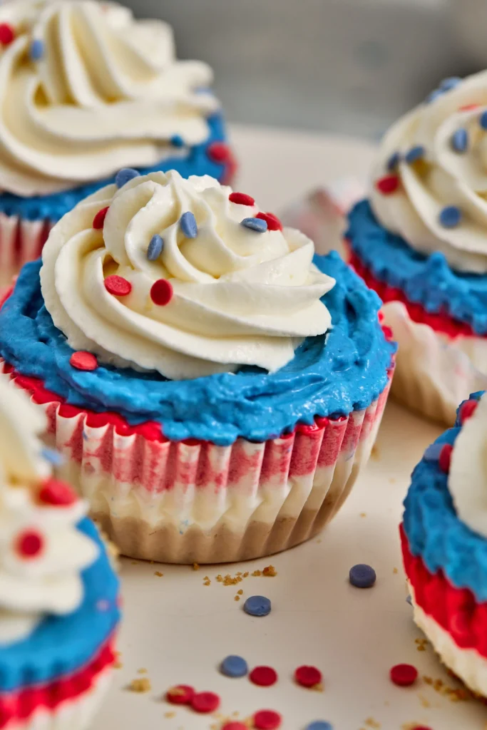 Close-up of red, white, and blue cheesecakes with white frosting, blue frosting accents, and red sprinkles on a white surface.