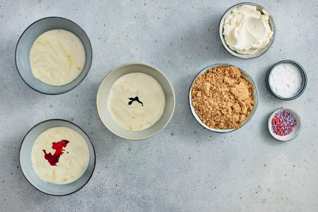 Top-view of several bowls with cream cheese filling and toppings (crumble, whipped cream) and a small sprinkle dish on a gray surface.