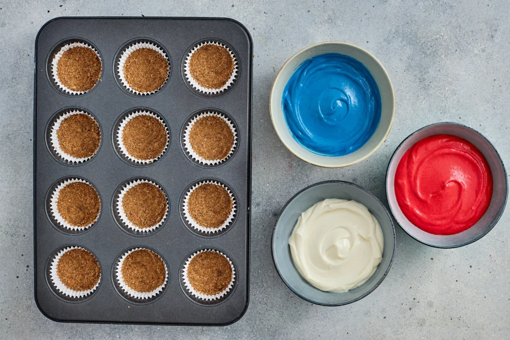 crust for the no-bake cheesecakes in a muffin tin with brown crumb topping, next to bowls of blue, red, and white frosting nearby.