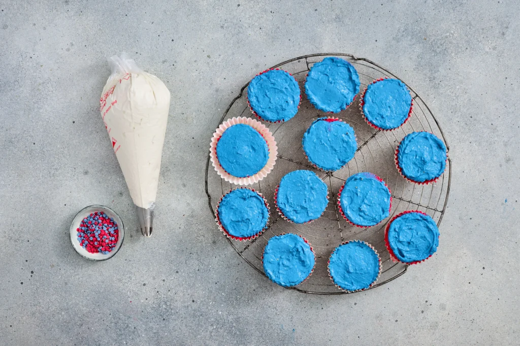 no bake cheesecakes with bright blue frosting arranged on a wire cooling rack, with a piping bag and a bowl of red-and-blue sprinkles nearby.