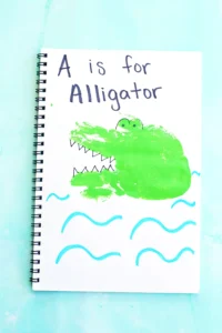 a letter A handprint craft for kids - a notebook with a green handprint stamped on it and decorated with look like an alligator with "A is for Alligator" written on top