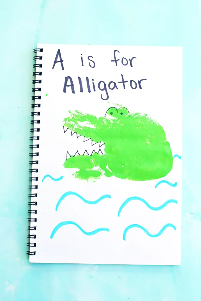 a letter A handprint craft for kids - a notebook with a green handprint stamped on it and decorated with look like an alligator with "A is for Alligator" written on top