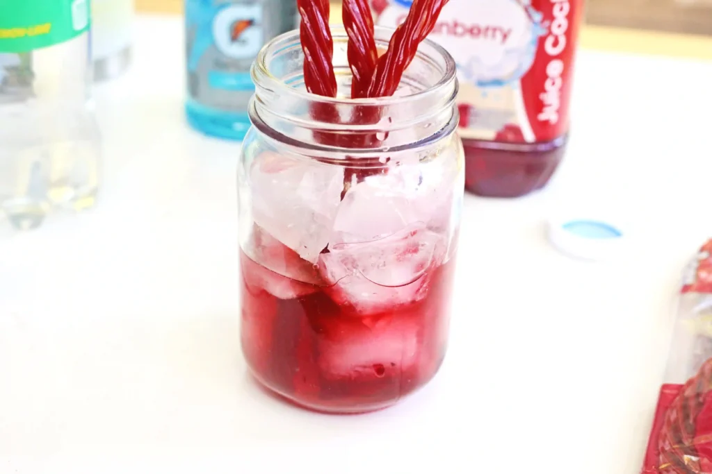 Close-up of a glass jar with red juice and ice, two red twizzlers, on a white surface with blurred bottles in the background