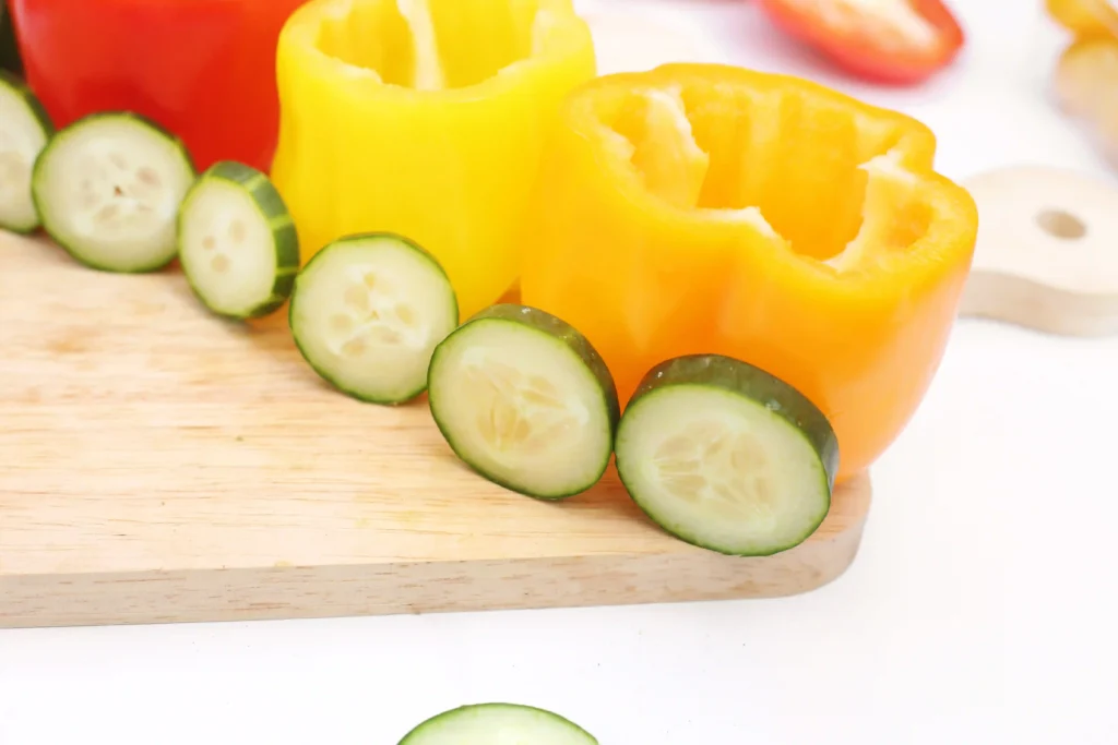 Sliced cucumber rounds and a yellow bell pepper on a wooden cutting board, with cucumbers