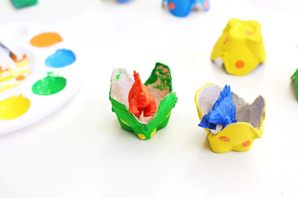Colorful painted egg-cartons with toy dinosaurs inside on a white surface with a paint plate nearby.