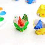 Colorful painted egg-cartons with toy dinosaurs inside on a white surface with a paint plate nearby.