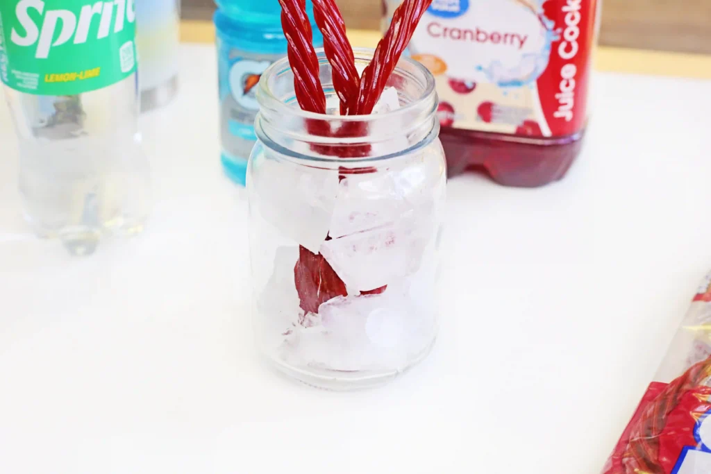 Mason jar filled with ice and red twizzlers, with colorful beverage bottles blurred in the background on a white surface.