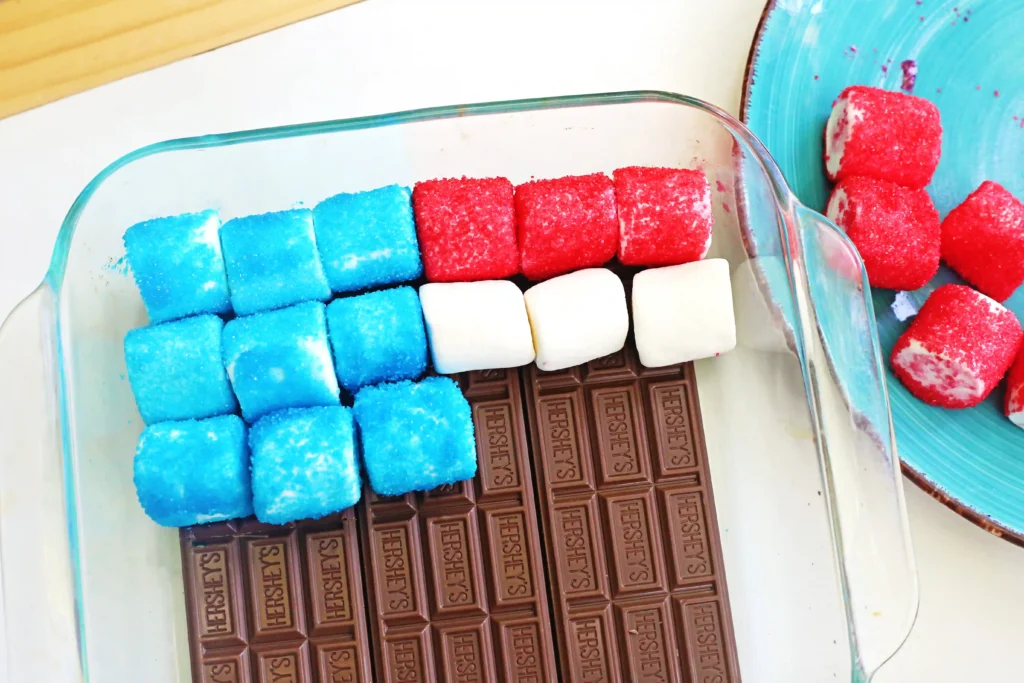 Blue and red sugar-coated marshmallows arranged in a glass dish above chocolate bars, with red-coated marshmallows on a turquoise plate nearby
