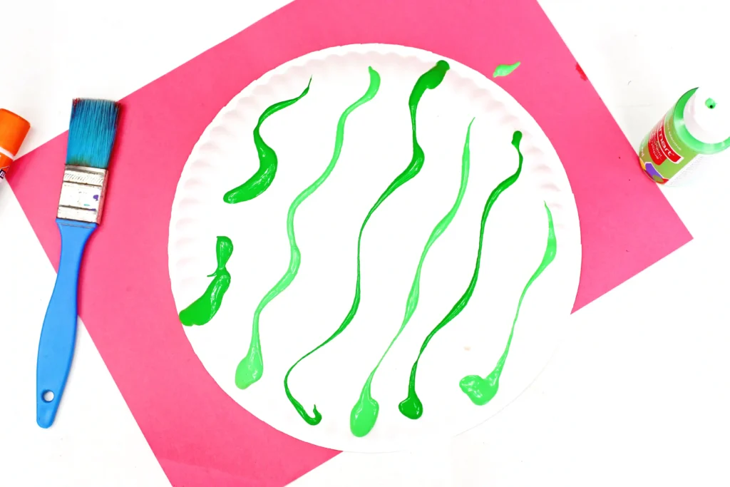 White paper plate with bright green paint squiggles, blue paintbrush, and pink background—an abstract painting setup.