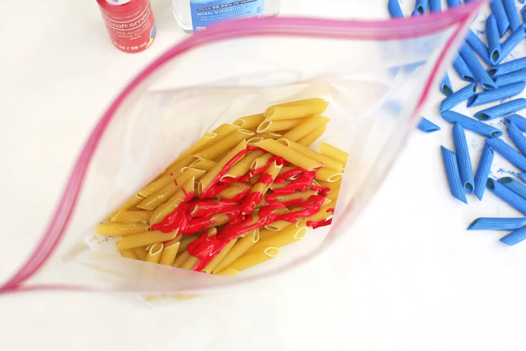 Patriotic Pasta Necklaces - Cultivating Brilliant Minds Bag containing uncooked penne pasta with red paint, blue pasta pieces scattered nearby on a white surface with art supplies in the background.