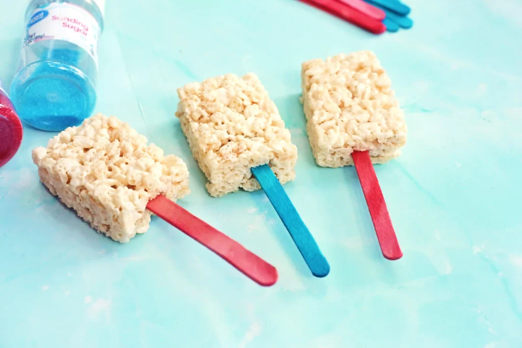 Patriotic Rice Krispie Pops - Cultivating Brilliant Minds Rice Krispies cereal bars on red and blue sticks laid on a turquoise surface, with a blue drink bottle in the background.