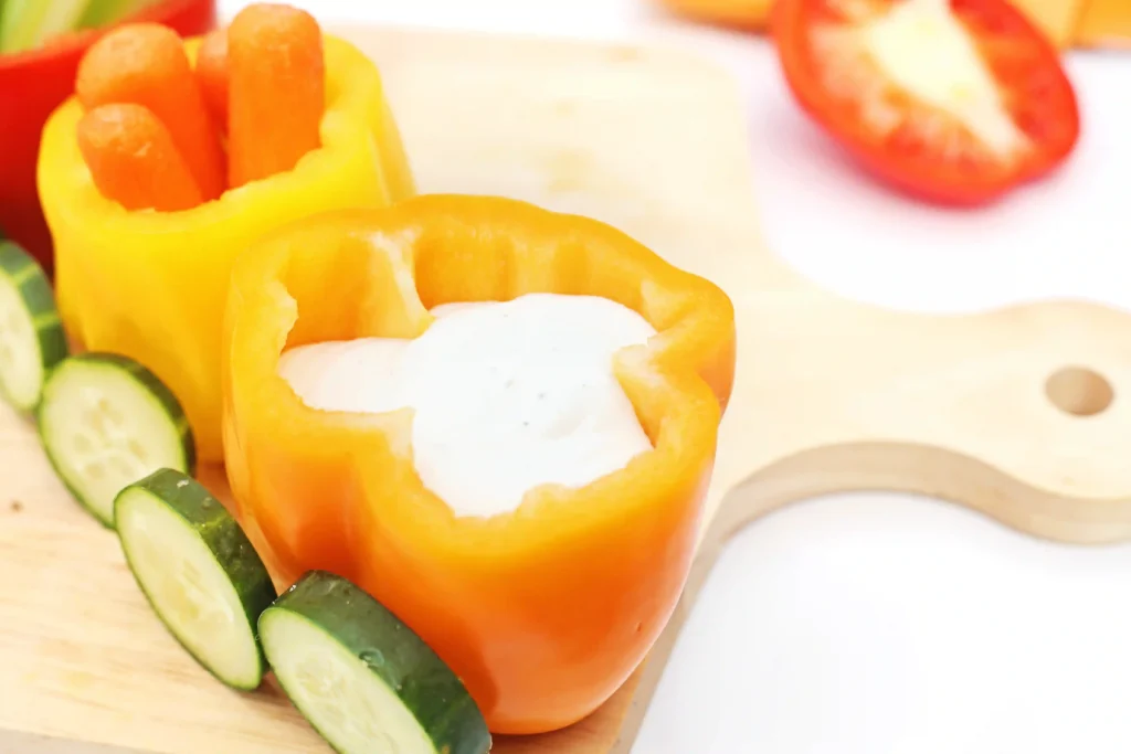 Orange bell pepper half-filled with ranch, surrounded by cucumber sticks on a wooden board