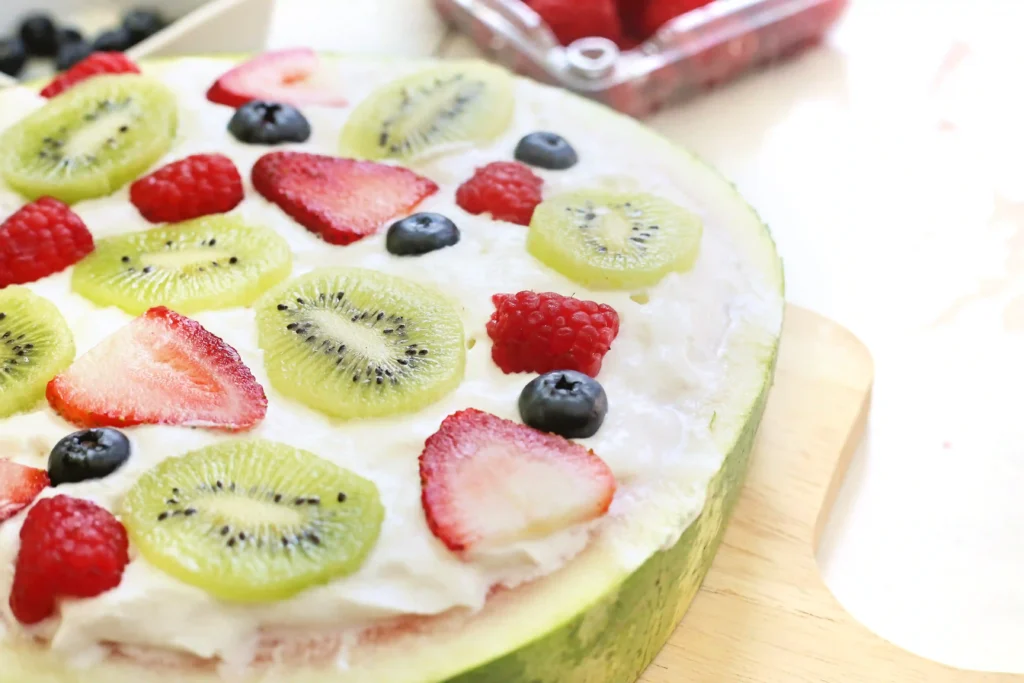 Fruit-topped watermelon with kiwi slices, raspberries, and blueberries on white frosting.