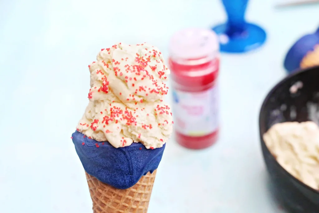 Ice cream cone with two scoops of vanilla ice cream covered in red sprinkles in a blue dipped waffle cone, held up close to the camera.