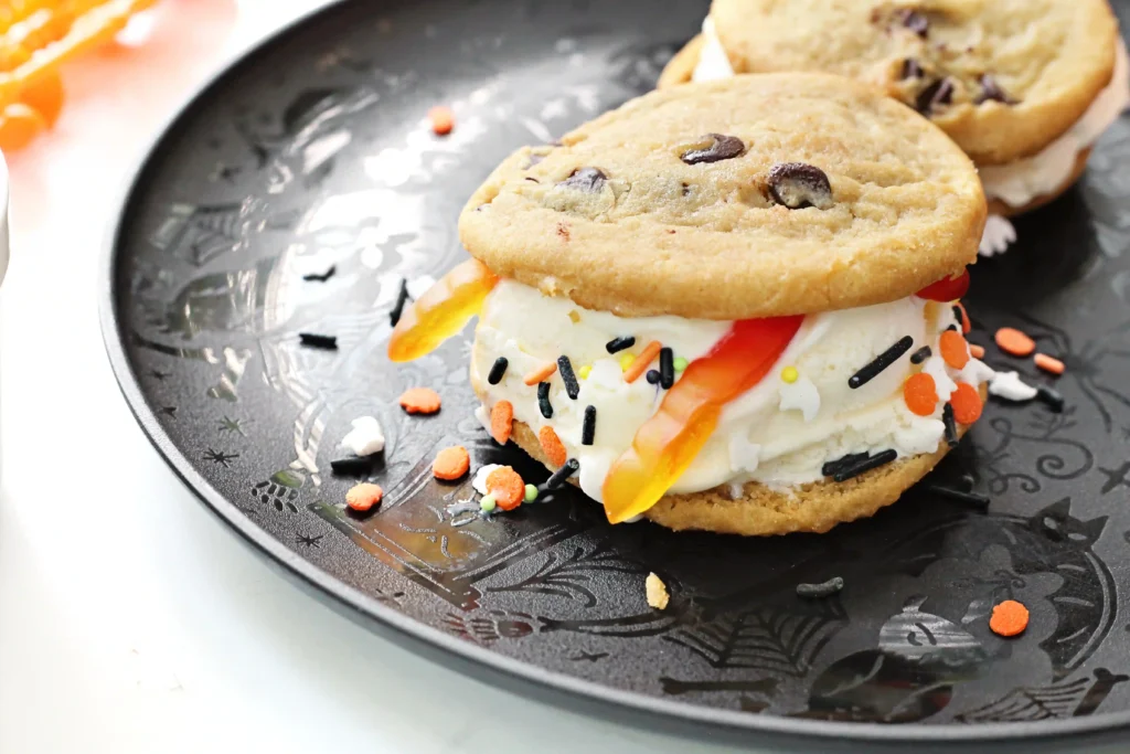 Close-up of a cookie sandwich with vanilla ice cream and Halloween-colored sprinkles on a dark decorative plate.