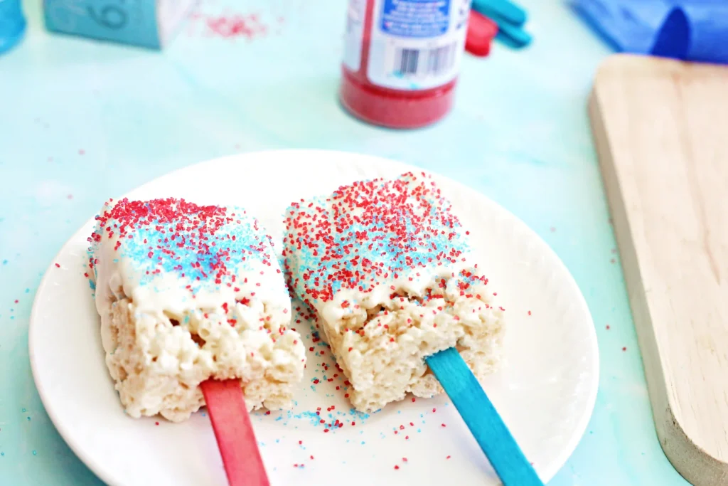 Patriotic Rice Krispie Pops - Cultivating Brilliant Minds rice krispie treats on red and blue stick with melted white chocolate on top and red and blue sprinkles