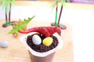 a dinosaur dirt cup that has chocolate pudding with crushed oreos, robins eggs, and a toy dinosaur on top