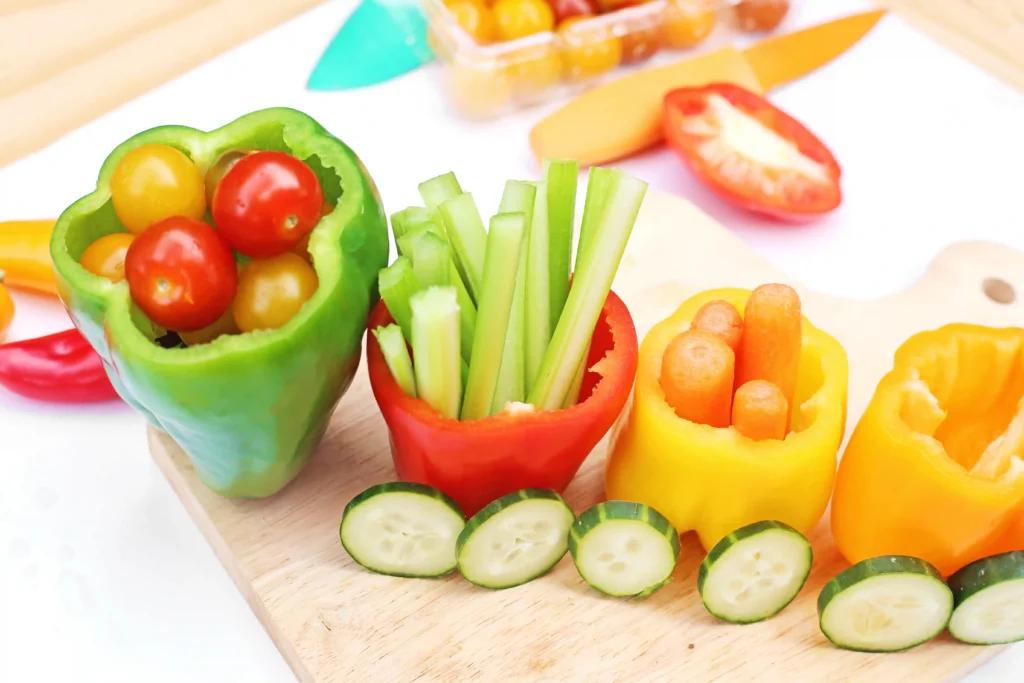 Colorful veggie snack platter on a cutting board: bell pepper cups filled with cherry tomatoes, cucumber rounds, celery sticks, carrot sticks, and yellow pepper slices.