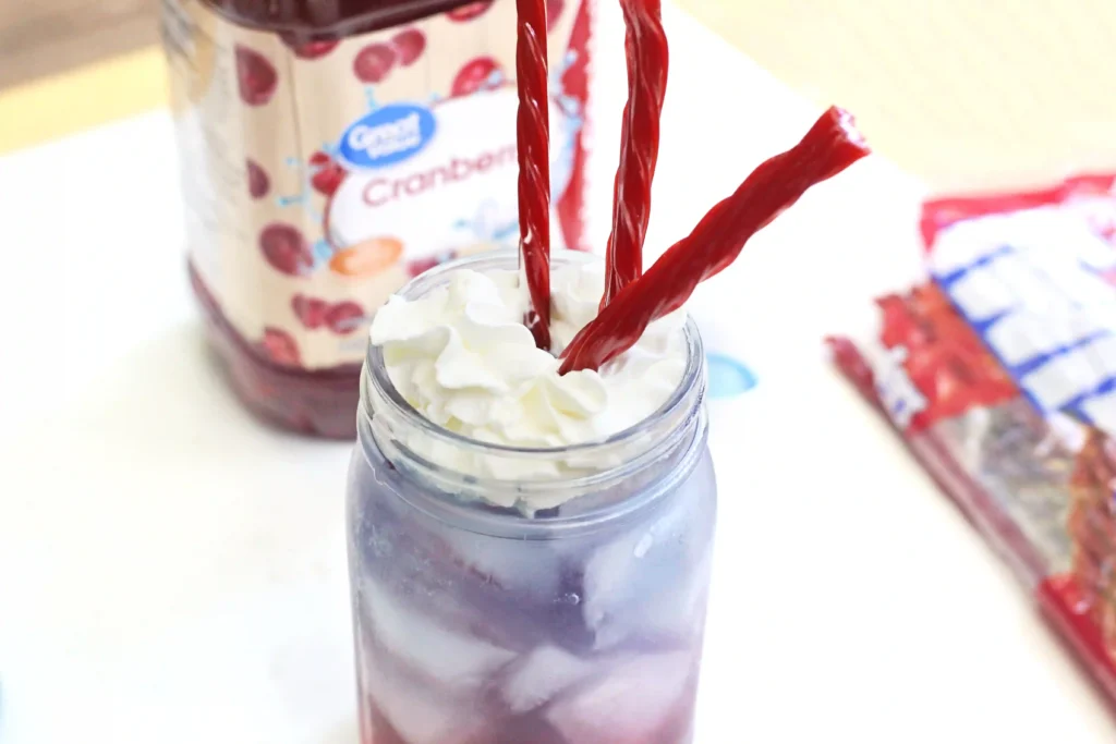 Close-up of a glass bottle topped with whipped cream and two red twizzlers, with a cranberry-flavored drink inside. This looks like a dessert beverage.