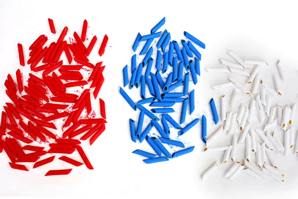 Patriotic Pasta Necklaces - Cultivating Brilliant Minds Red, blue, and white short pasta pieces scattered in three clusters on a white surface.