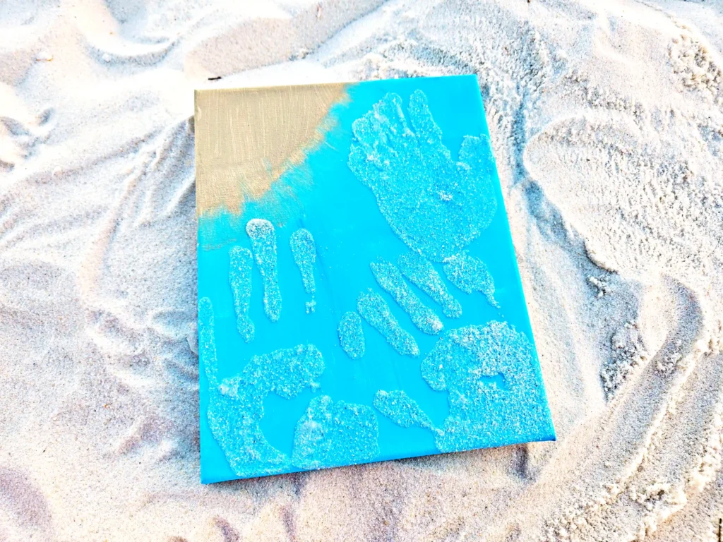 Blue canvas lying on sandy beach with sand pressed into blue-painted handprints and shapes on its surface.