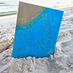 Blue art board stuck in sandy beach with turquoise paint drips and ocean in the background