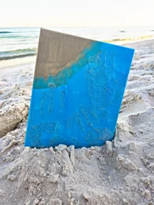 Blue art board stuck in sandy beach with turquoise paint drips and ocean in the background