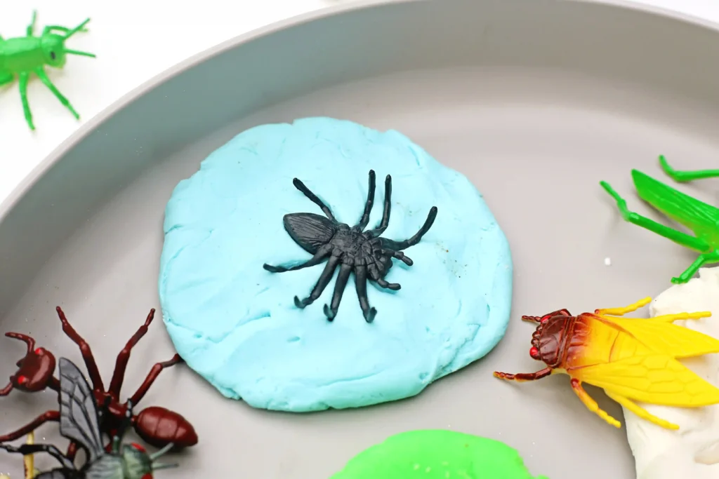 Blue playdoughwith a black plastic bug on top, surrounded by colorful craft insects on a plate.