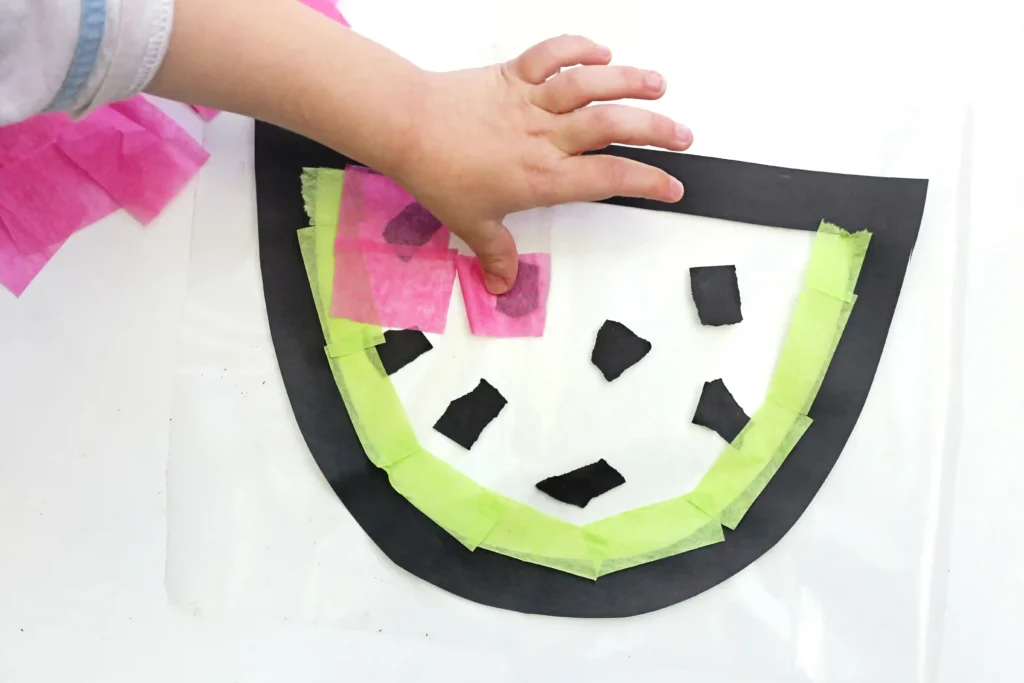 Child's hand arranging pink and lime tissue paper cutouts on a black circular art piece for a collage project.
