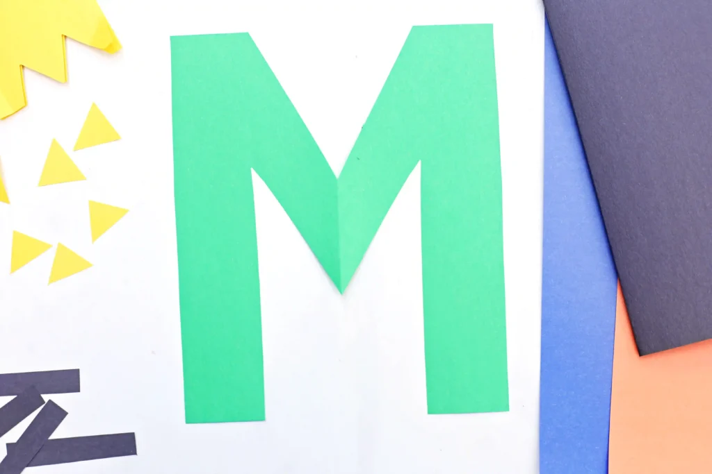 Close-up of a large green letter M on a white background with yellow triangular accents on the left and blue shapes on the right.
