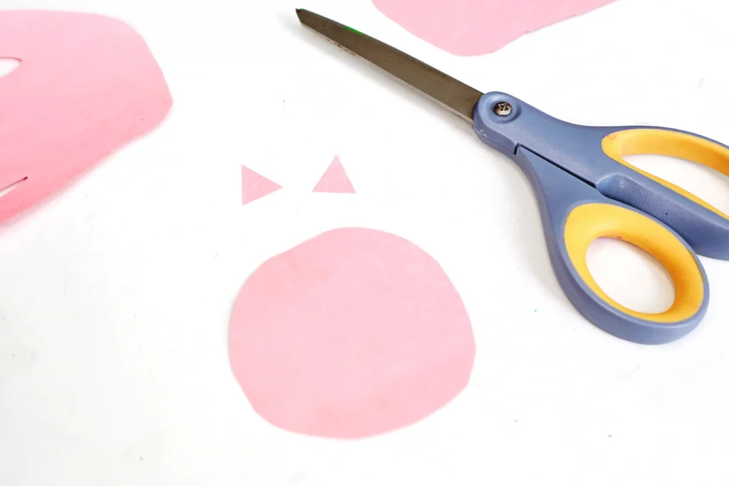 Pair of gray and yellow scissors on a white surface beside pink crafting paper shapes.