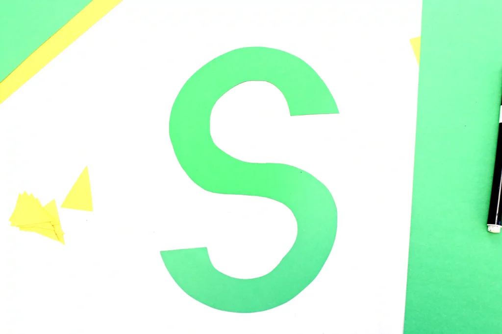 Close-up of a large green letter S on a white poster with a lime-green border and yellow corner shapes.