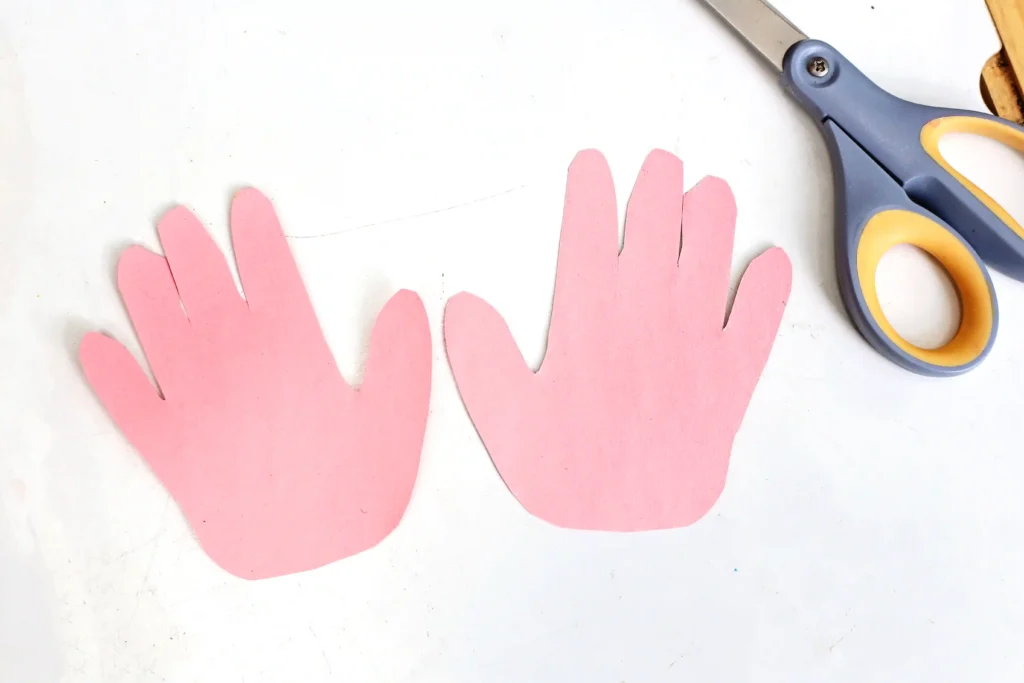 Pink paper hand cutouts arranged on a white surface with a pair of scissors on the right.