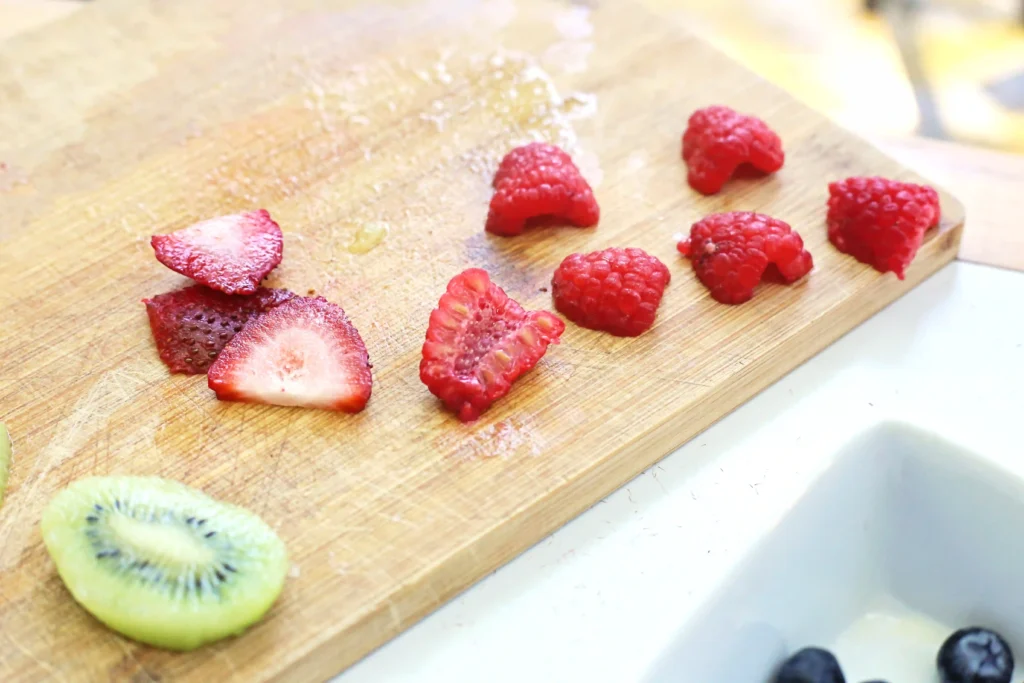 Sliced strawberries, raspberries, and kiwi on a wooden cutting board, with blueberries at the edge and bright kitchen light behind.