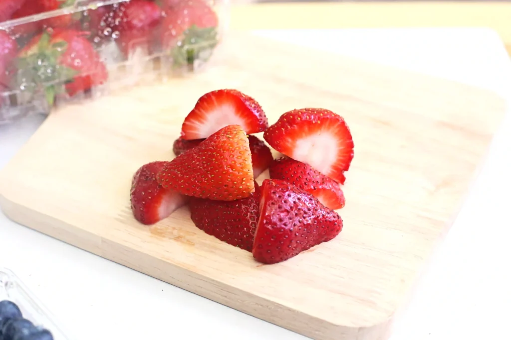 Sliced strawberries on a light wooden cutting board, close-up view with a blurred strawberry bouquet in the background
