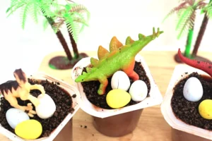 a dinosaur dirt cup that has chocolate pudding with crushed oreos, robins eggs, and a toy dinosaur on top