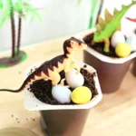 Tiny dinosaur figurine perched on chocolate soil in a small cup with pastel blue and yellow candy eggs, against a background of potted plants and a wooden surface.