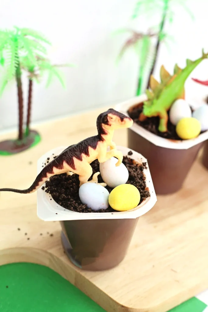 Tiny dinosaur figurine perched on chocolate soil in a small cup with pastel blue and yellow candy eggs, against a background of potted plants and a wooden surface.