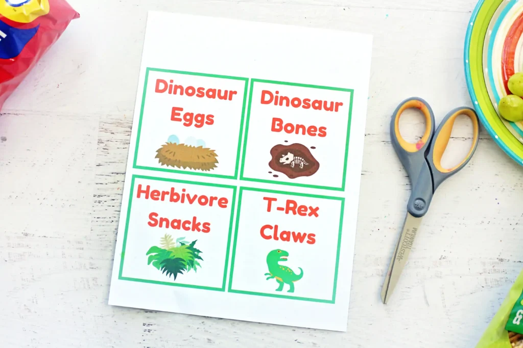 Printable Food Labels  'Dinosaur Eggs' and 'Dinosaur Bones' with illustrations of an egg and a fossil, on a white table near scissors and plates.