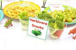 Colorful snack display with broccoli florets and celery sticks in clear cups, labeled Herbivore Snacks for kids' party.