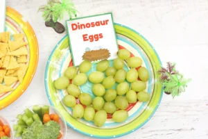 Colorful party spread with green grapes and crackers, and a sign reading Dinosaur Eggs beside a dinosaur egg illustration.