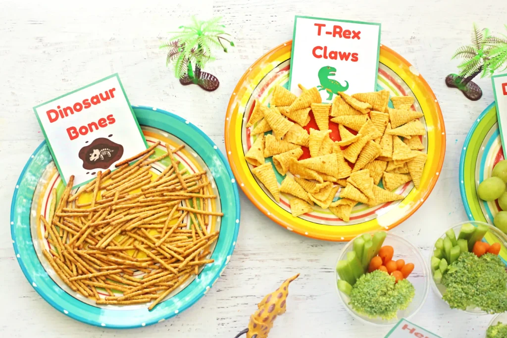 Party snack table with dinosaur-themed signs: Dinosaur Bones and T-Rex Claws next to plates of pretzel sticks and cheese crackers.