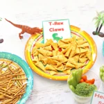 Bright plate of triangular corn chips labeled 'T-Rex Claws' with a small green dinosaur nearby.
