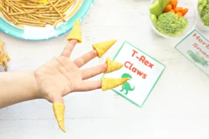 Hand wearing small corn-cone “claws” on fingertips, with a plate of fries and a veggie tray in the background