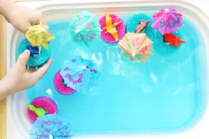 a bin filled with blue water and pool noodle slices with toy dinosaurs and mini umbrellas