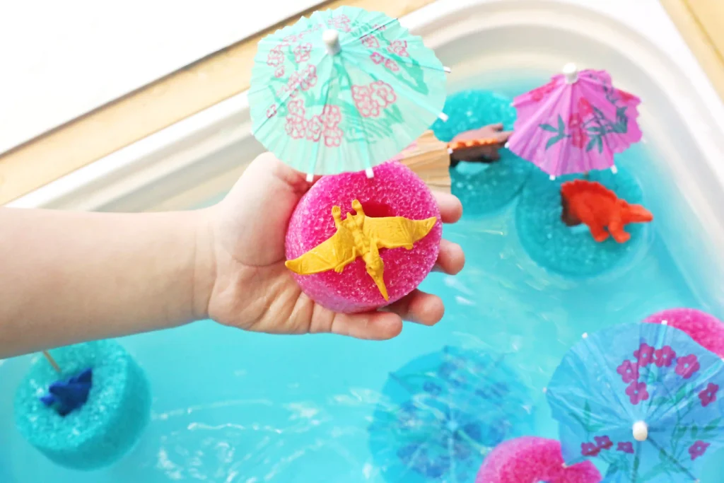 Child's hand holds a bright pink sponge with a small yellow dragon on it, with colorful paper umbrellas in a turquoise bath scene in the background.